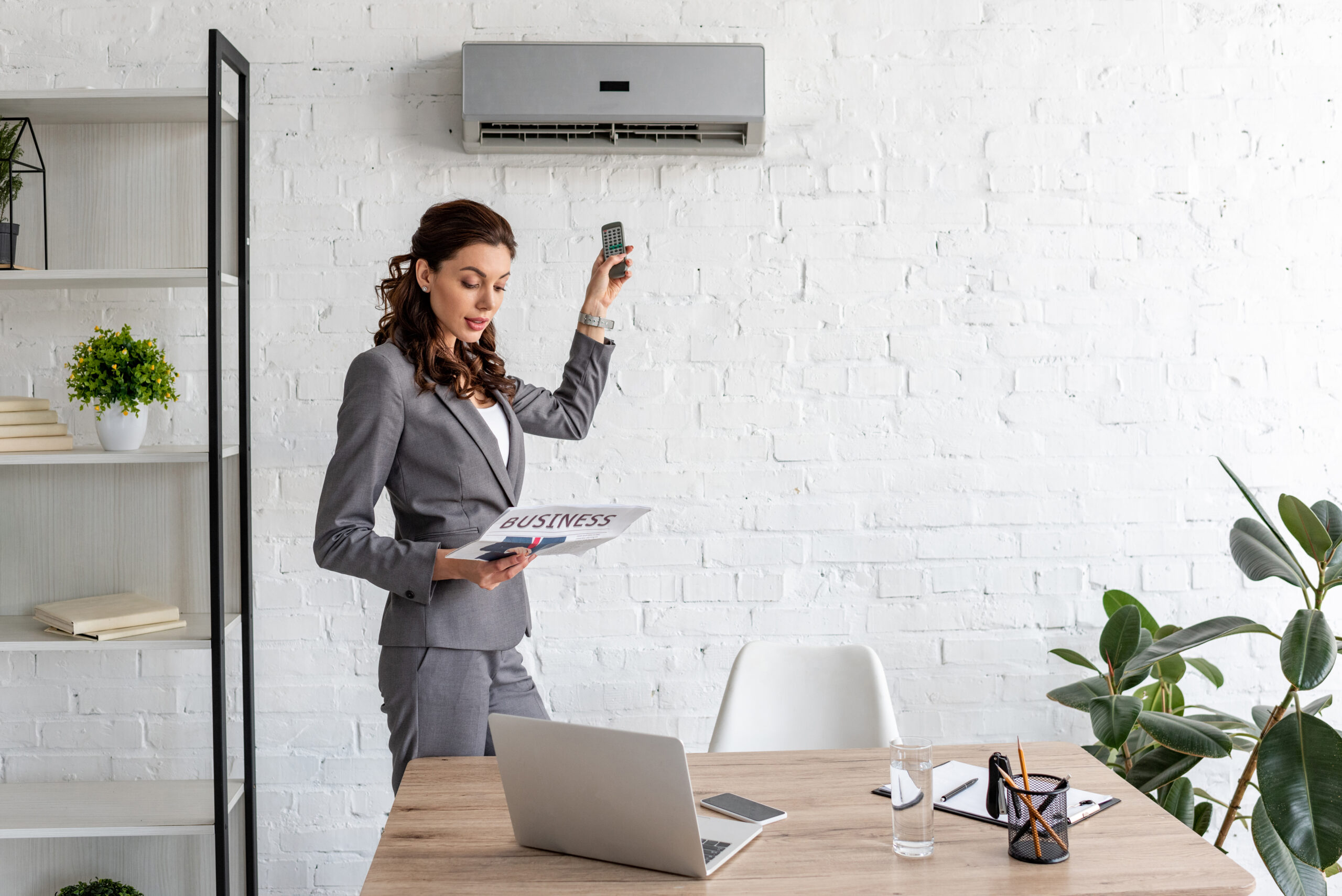 beautiful businesswoman reading business newspaper while standing under air conditioner with remote